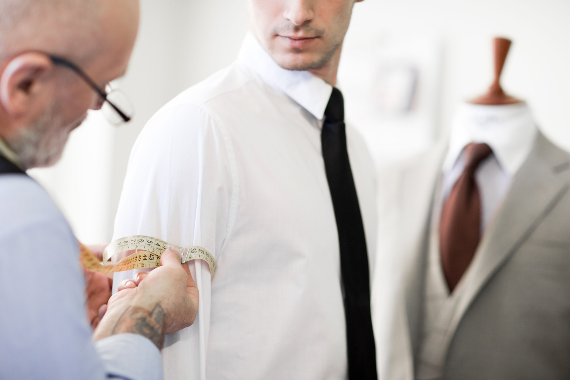 Close-up of busy tailor measuring arm of businessman in white shirt looking at tape measure in sewing studio