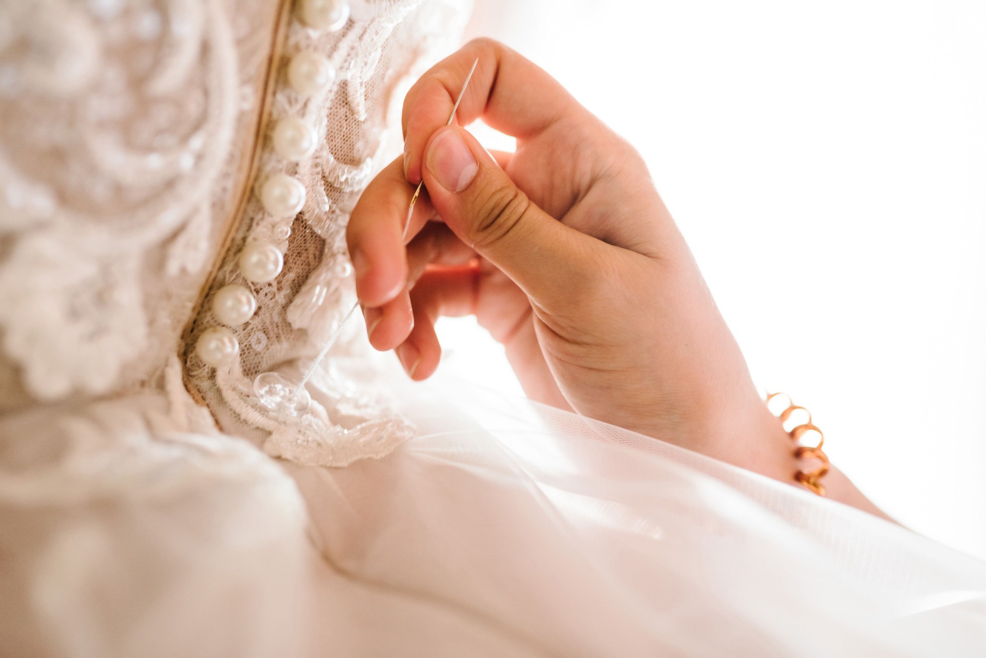 Close-up of work process of a tailor in her studio. Clothes shop for wedding dresses. Designer hands sewing wedding dress. Hands of woman sew beads and white lace on mannequin or bride.