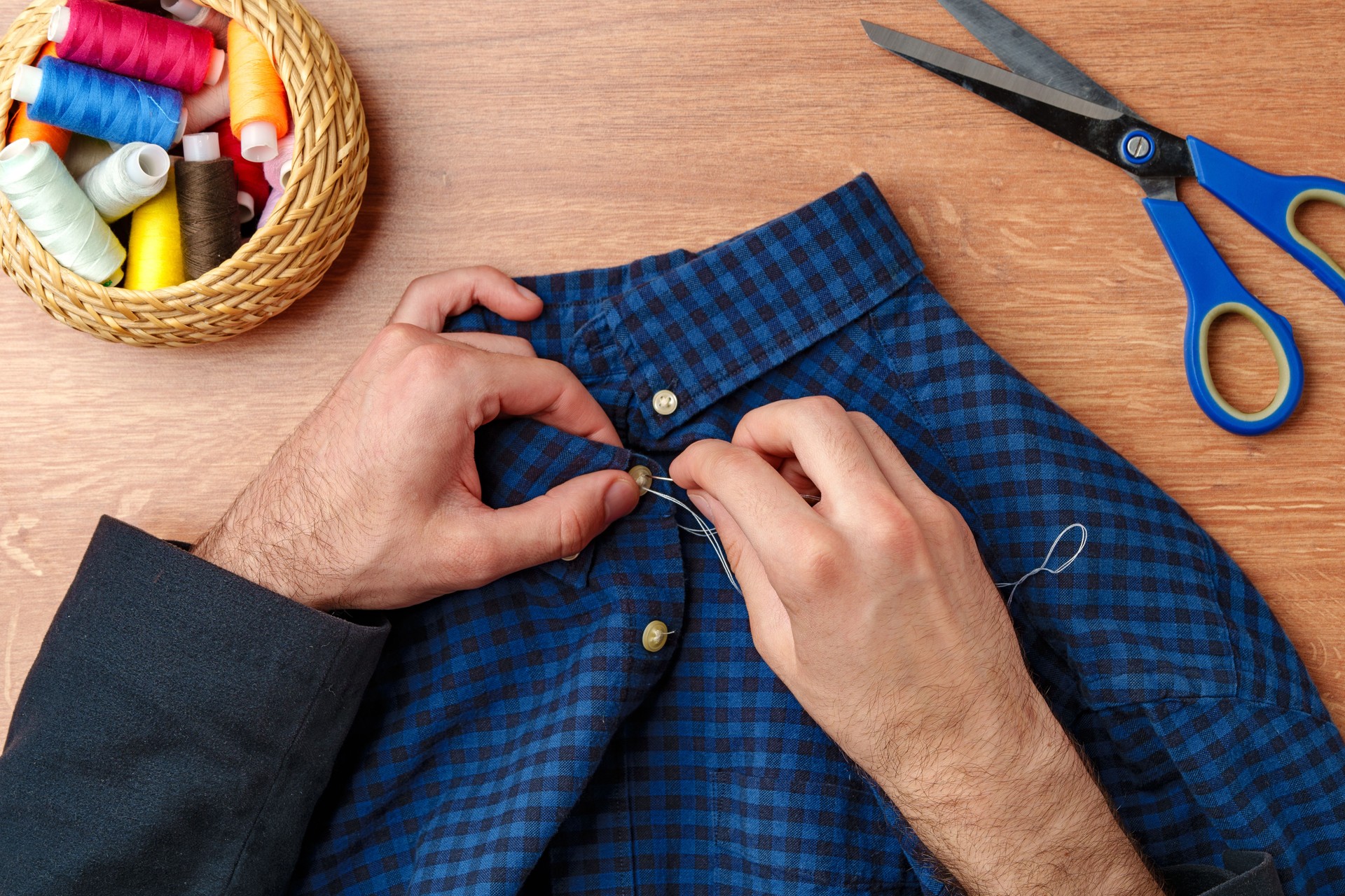 A person sewing a button on a blue plaid shirt with sewing tools and threads on a wooden table